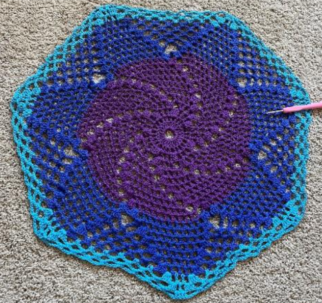 Proyecto de tapete de crochet terminado que muestra el patrón en espiral que comprende púrpura en el centro, rodeado por una capa azul oscuro y una capa azul claro alrededor del exterior.
