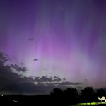 purple and green aurora lights filling the night sky with silhouettes of a few buildings at the bottom